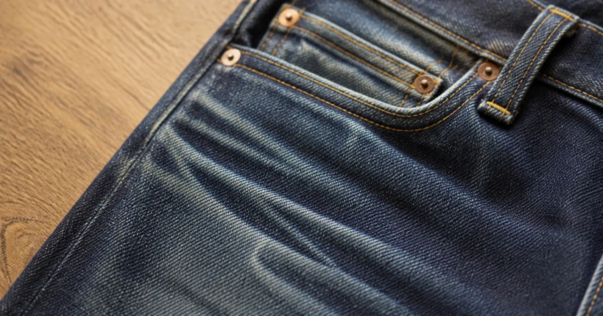 A close-up of the front pocket area of well-worn raw denim showing developing whisker and honeycomb fade patterns where the fabric bends, with the indigo lightening to a softer blue in the creases on a matte oak desk in soft natural daylight (AI generated illustration)