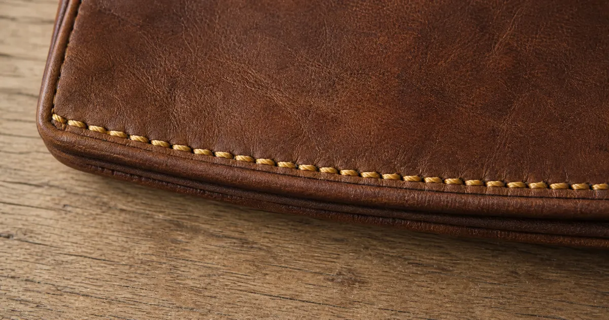 A close-up of the stitching along the edge of a vintage leather handbag, showing tight, evenly spaced stitches in a slightly mustard-yellow thread, all running parallel to the leather edge on a matte oak desk in soft natural daylight (AI generated illustration)